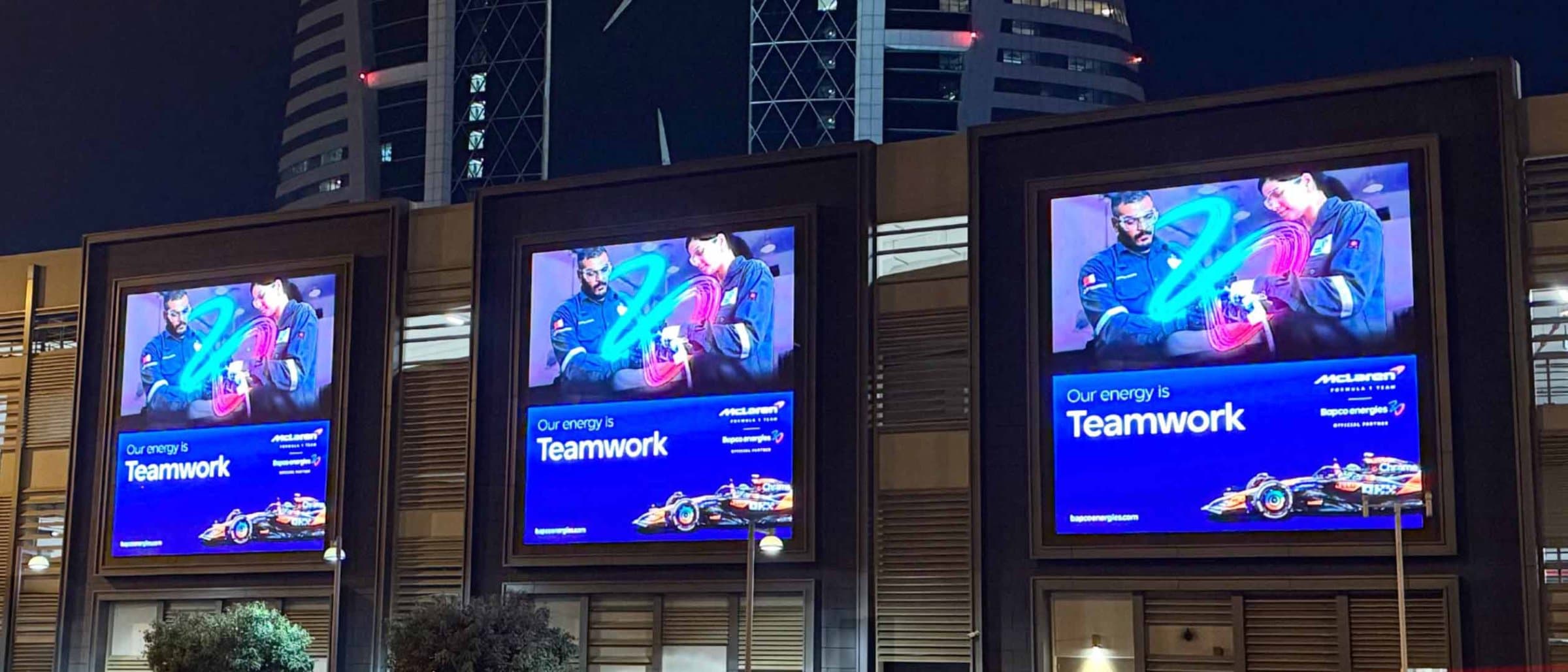 Digital screens on a building facade showing the Bapco Energies McLaren partnership and the line 'Our energy is Teamwork'