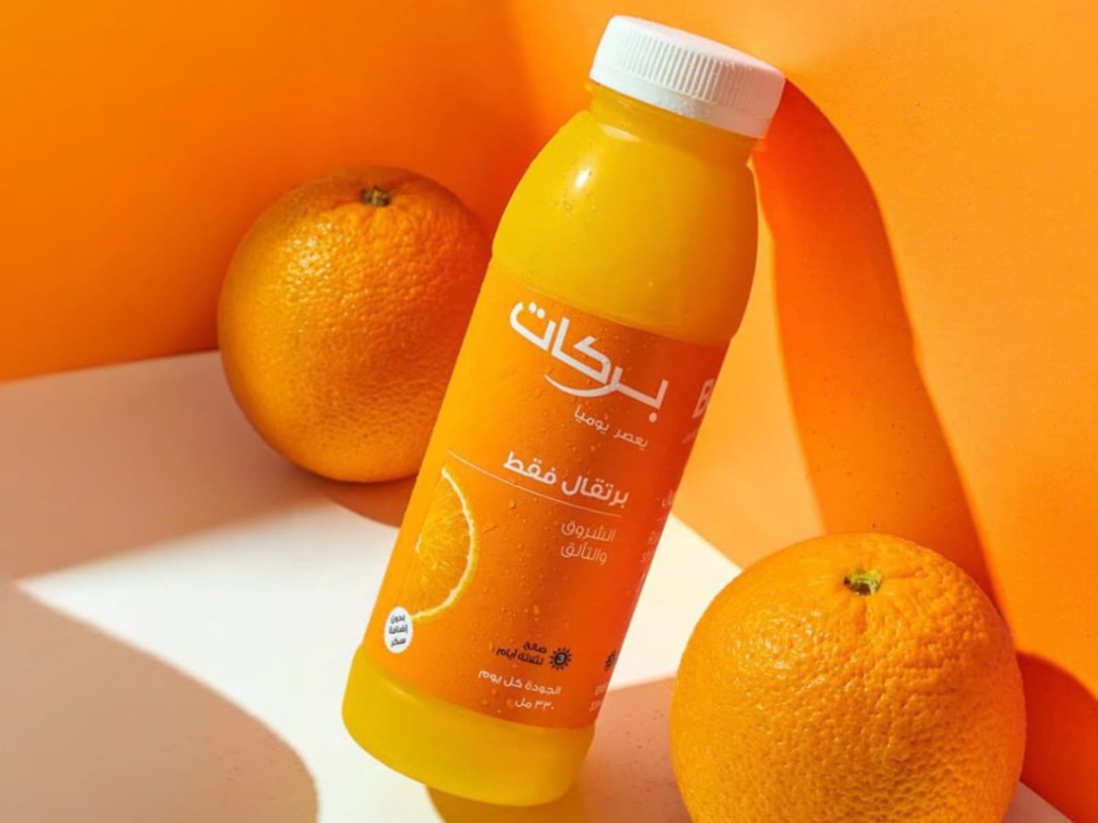 Two Barakat juice bottles styled with fresh fruit - orange juice against an orange background, pomegranate juice against red